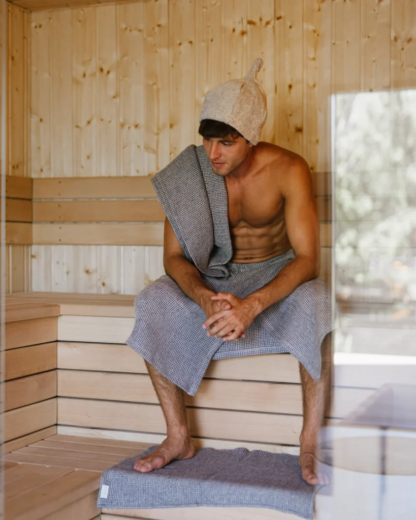 mężczyzna siedzący w saunie, owinięty kiltem do sauny wafel natural jeans blue i ręcznikiem do sauny, ubrany w czapkę do sauny